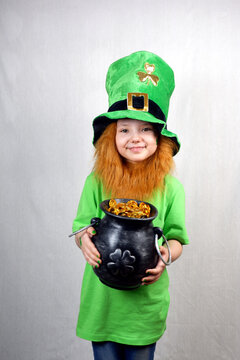 Saint Patricks Day Holiday. Pretty Smiling Small Girl In Green Clothes With Decorative Red Beard And Leprechaun Hat, Green Shamrock Leaf On Her Cheek, Showing Off Cast Iron Lepreaun Pot, Full Of Gold