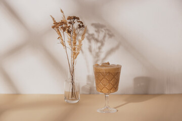 Coffee drink in a stemmed glass with a bouquet of dried flowers on a beige and white neutral background with shadows from the summer sun. Background with copy space