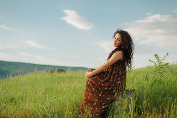 Lifestyle shot of curly hair girl sitting on stone in field and looking at camera