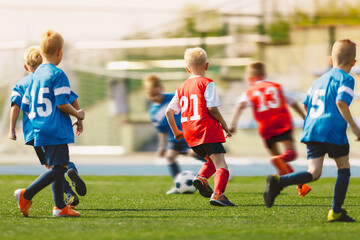 Young Footballers Kicking Football Match.Football Soccer Players Running with Ball. Soccer Players Running After the Ball. Kids in Soccer Red and Blue Uniforms. Soccer Stadium in the Background