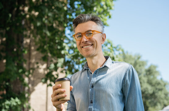 Smiling mature businessman drinking coffee outdoors. Happy handsome man wearing stylish eyeglasses holding cup of tea, looking at camera. Coffee break concept	
