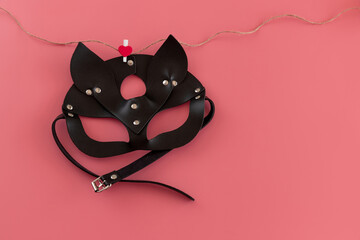 Black leather sexy cat mask hung on clothespin with hearts on a string on pink background with space for text. Concept of Valentine's Day. Copy space, top view. The concept of Valentine's Day.