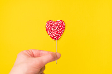 Handholds red heart-shaped lollipop candy isolated on an empty colorful yellow background. Symbol of love for Happy Women's, Valentine's Day, Birthday greeting romantic postcard. Flat lay, top view
