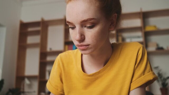 A close-up view of a serious young redhead woman is writing while thinking about something sitting at the table in the office