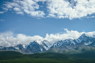 Fototapeta premium Awesome mountains landscape with sunlit high snowy pinnacle among low clouds in blue sky. Atmospheric highland scenery with snow-white big mountain top in sunlight. Wonderful snowy mountain range.