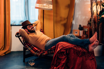 home relax. country style american man wearing a cowboy hat lying in a chair in a comfort room and drinking a glass of wine
