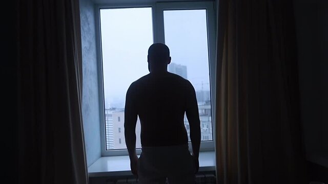 Young man silhouette opens curtains standing at large window against city buildings slow backside motion