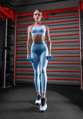 Beautiful tall blonde posing in the gym with dumbbells in her hands against the background of the...