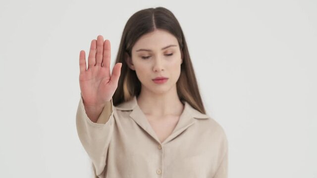 A serious young woman is showing stop gesture standing isolated over white background in stud