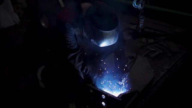 A laborious worker in a uniform and a protection mask during welding process. Clip. Welder in a workshop of a plant working with metal details.