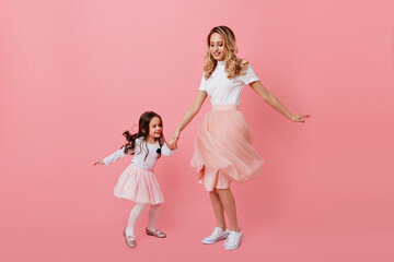 Positive blonde mom and dark-haired daughter in lush romantic skirts are dancing boogie woogie on pink background