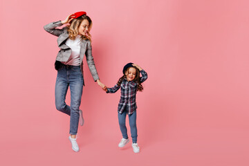 Adult curly blonde woman and her female child are dancing on pink background. Portrait of mother and daughter in checkered jackets, jeans and berets