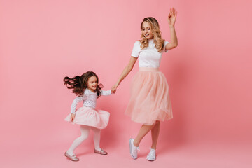 Full-length shot of cheerful adult lady and little girl in light dresses dancing on isolated background