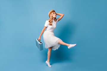 Full-length picture of traveler girl running with hand suitcase. Lady in white fluffy skirt, t-shirt and sneakers posing on isolated background