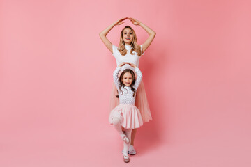 Mom and daughter pose as ballerinas on pink background. Portrait of curly adult blonde woman in white T-shirt and her younger sister in tutu