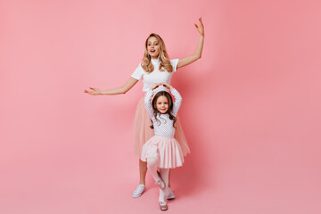 Adult lady and little girl in pink skirts and light tops dance ballet on pink background