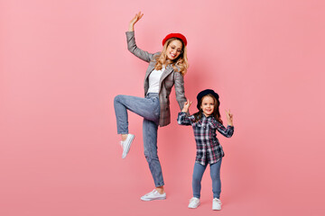 Studio portrait of cheerful adult and little girls in jeans, stylish shirts and berets having fun on pink background
