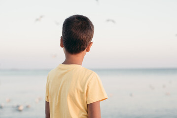 boy in a yellow shirt looks far away to horizon of sea standing alone on beach at sunset. View from...