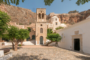 Church tower on sunny village square with some trees and an old fortress on top of the hills in the middle of Monemvasia, Peleponnese, Greece