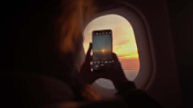 Passenger taking a mobile photography of an intense sunset through the airplane window. The phone is in the hands of a girl out of focus, photographing the sky and the wing of the plane