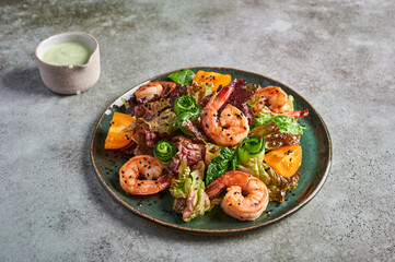 Homemade salad with fried shrimp, yellow tomatoes, arugula, spinach, lettuce and sauce on a ceramic plate on texture background, close up, copy space