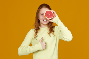 adorable redhead little child girl holding fresh juicy grapefruit and showing Ok gesture isolated on yellow background
