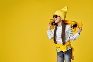 smiling little child girl in hat and sunglasses posing with yellow skateboard on yellow background. copy space