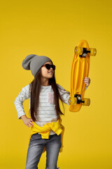 cool little child girl in hat and sunglasses holding yellow skateboard on yellow background.