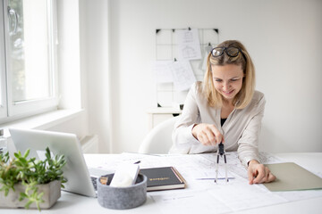 beautiful, young and blond female architect with glasses is drawing something with a compass in a plan in her office and is happy