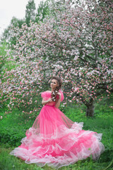 Fototapeta premium A beautiful young girl with long hair in a light pink ball gown walks through a blooming apple orchard