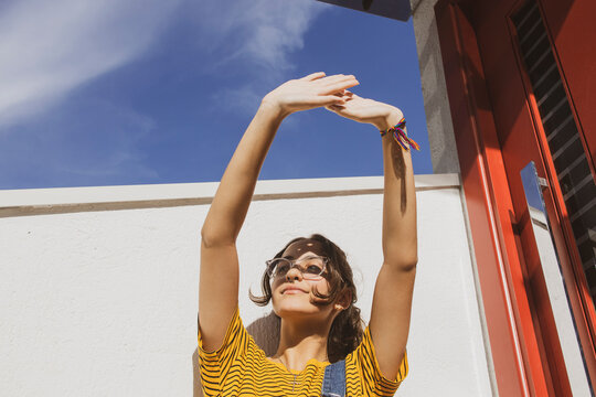 Portrait of a teenager girl wearing clear transparent glasses and colorful clothes against white wall protecting eyes from sunlight with her hand.	
