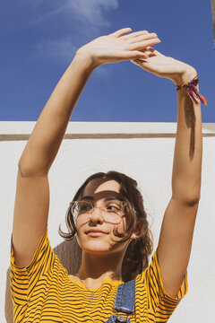Portrait of a teenager girl wearing clear transparent glasses and colorful clothes against white wall protecting eyes from sunlight with her hand.	