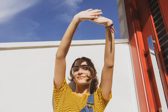 Portrait of a teenager girl wearing clear transparent glasses and colorful clothes against white wall protecting eyes from sunlight with her hand.	