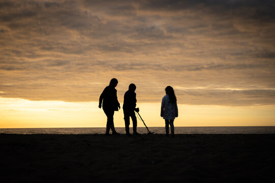Family children on holiday vacation silhouetted treasure detecting at sunset beach near ocean metal detector and amazing orange dramatic sun sky over sea 