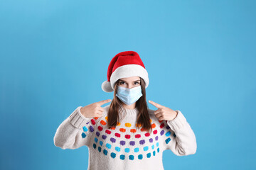 Fototapeta na wymiar Pretty woman in Santa hat and medical mask on light blue background