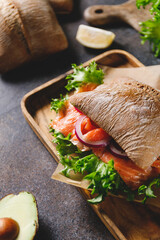 Healthy sandwich with rye bread bun, salmon, avocado, onion and salad served on a wooden rustic board.