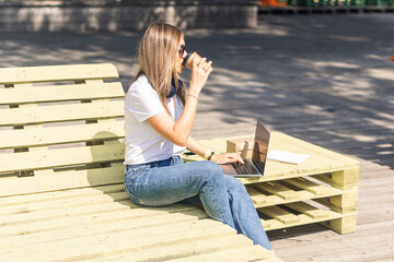 Beautiful woman working at notebook sit down on bench outside and drinking coffee. Using computer and online shops. Happy lady girl with light hair. Distance learning online education.