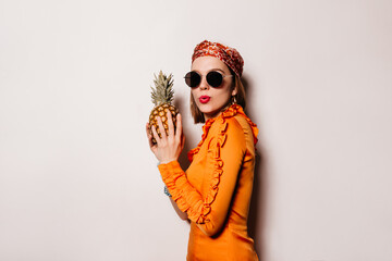 Young woman with red lips is holding pineapple. Portrait of naughty girl in orange outfit and...