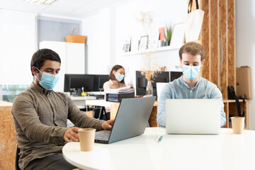Coworkers concentrated on their laptops and wearing protective face masks. Working in the office during Coronavirus pandemic concept.