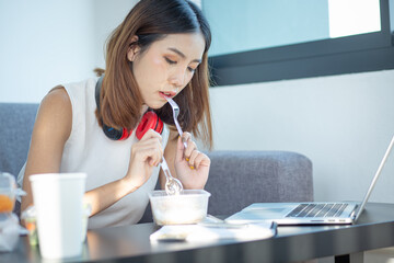 Young attractive asian female hungry eat doughnut take away snack food with full mouth look at computer notebook at home in busy work from home multitask unhealthy meal lifestyle concept.
