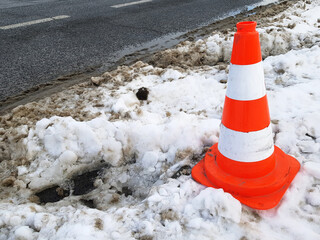 Bright orange traffic cone standing on dirty melting snow at roadside. After cleaning the road with...