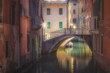 Bright, colourful Venetian architecture with a bridge over a calm canal and a lone boat during a quiet night in a secluded residential area of old town Venice, Italy.