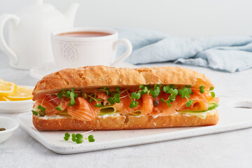 Big sandwiche with salmon, cream cheese, cucumber slices on white concrete table. Morning healthy breakfast with fish and cup of drink, side view, close up