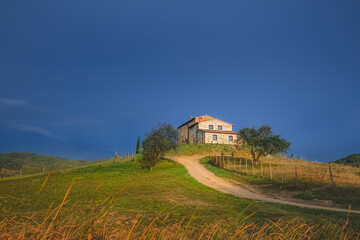 Obraz premium A lone countryside farmhouse in Radicondoli catches some golden sunset or sunrise light with a moody dark blue sky as a backdrop in Tuscany, Italy.