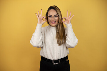 Pretty woman with long hair doing ok sign with fingers and smiling, excellent symbol against yellow wall