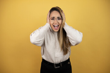 Pretty woman with long hair crazy and scared with hands on head, afraid and surprised of shock against yellow wall