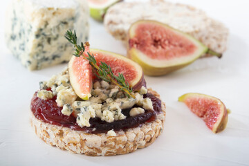 Puffed exploded wheat grains with slices of figs and blue cheese on a thin layer of jam on a light wooden table against a background of fruits and a cheese head. Close-up