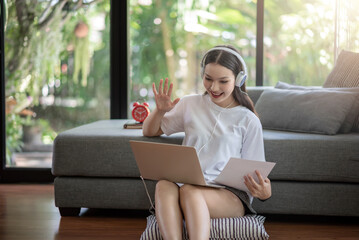 Asian woman happy with headphones look at the laptop screen greetings to a friend at home online.