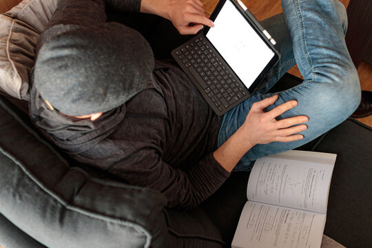 Hombre joven cauc&aacute;sico sentado en el sofa con las piernas cruzadas usando una tablet junto a un libro de texto mientras est&aacute; estudiando a dist&aacute;ncia desde casa.