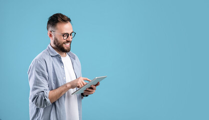 Smart millennial guy in glasses using tablet computer over blue studio background, banner design with copy space